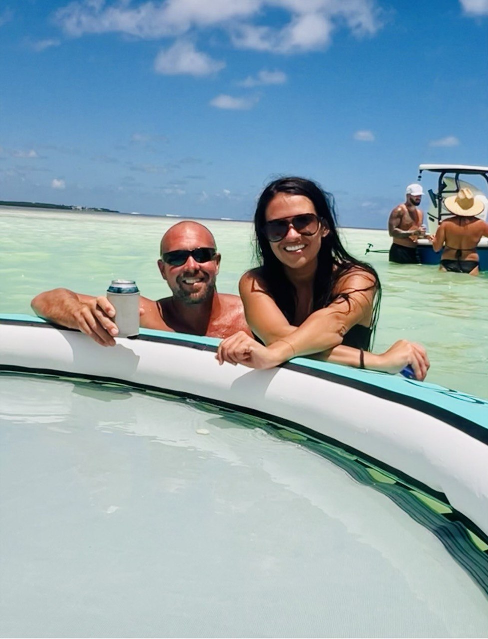 Couple smiling beside a floating lounge in shallow turquoise sandbar water